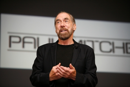 John Paul DeJoria, co-founder of Paul Mitchell hair products and The Patrón Spirits Company, standing confidently in front of a Paul Mitchell logo