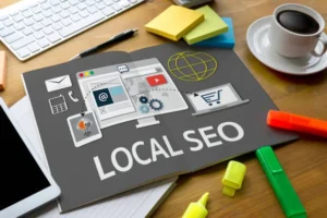 Overhead view of a wooden desk with a keyboard, coffee cup, and a black booklet open to a page titled LOCAL SEO showing icons for email, website, mapping, and a shopping cart