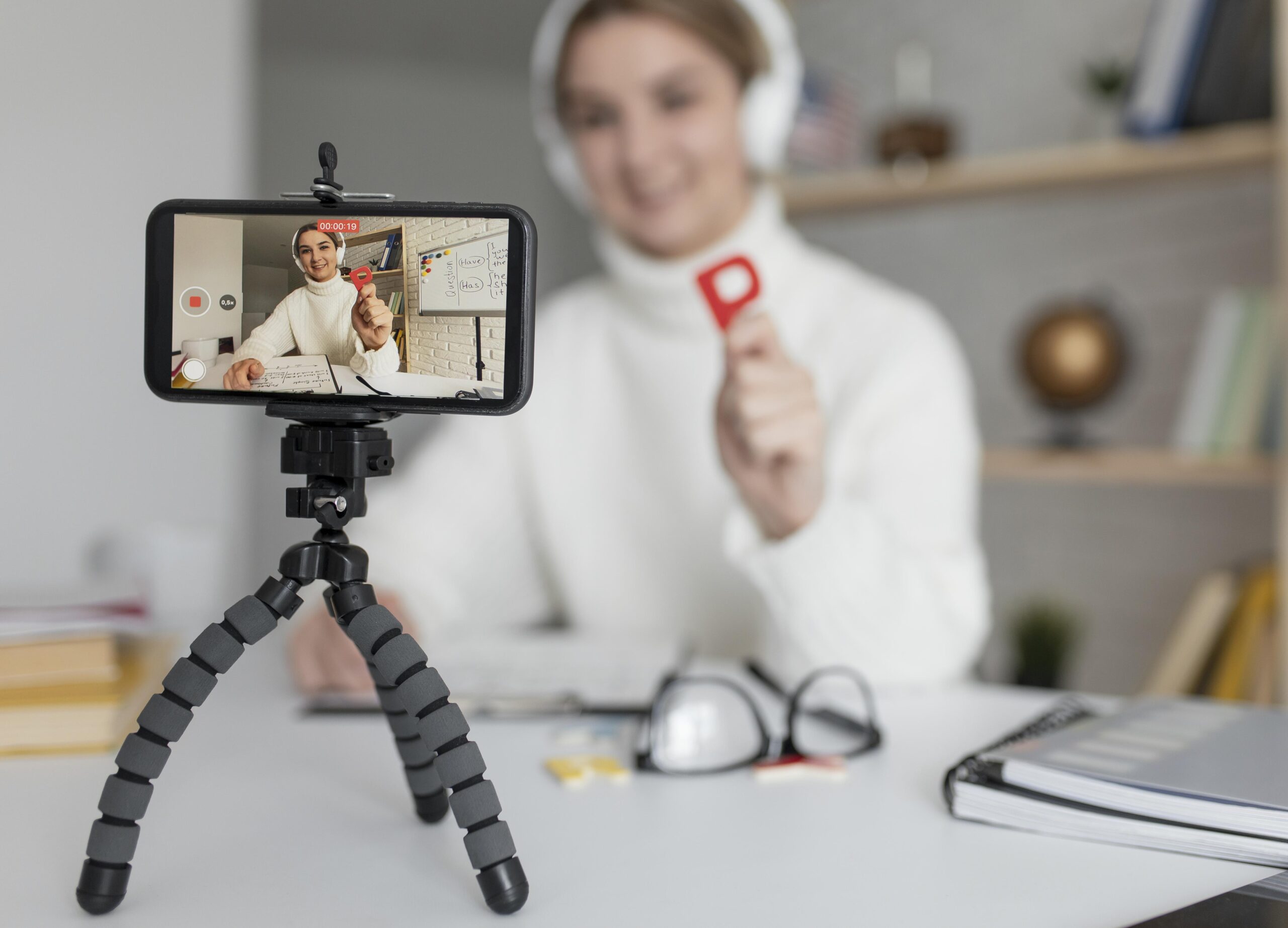 young-woman-teaching-kids-english-lesson-online Vlogger or educator creating professional video content at a desk with supplies, using a mobile phone camera to film a tutorial or online course