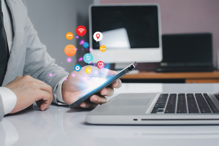 Person in a business suit using a mobile phone at a desk with a laptop, showing glowing icons for WiFi, mail, home, and shopping