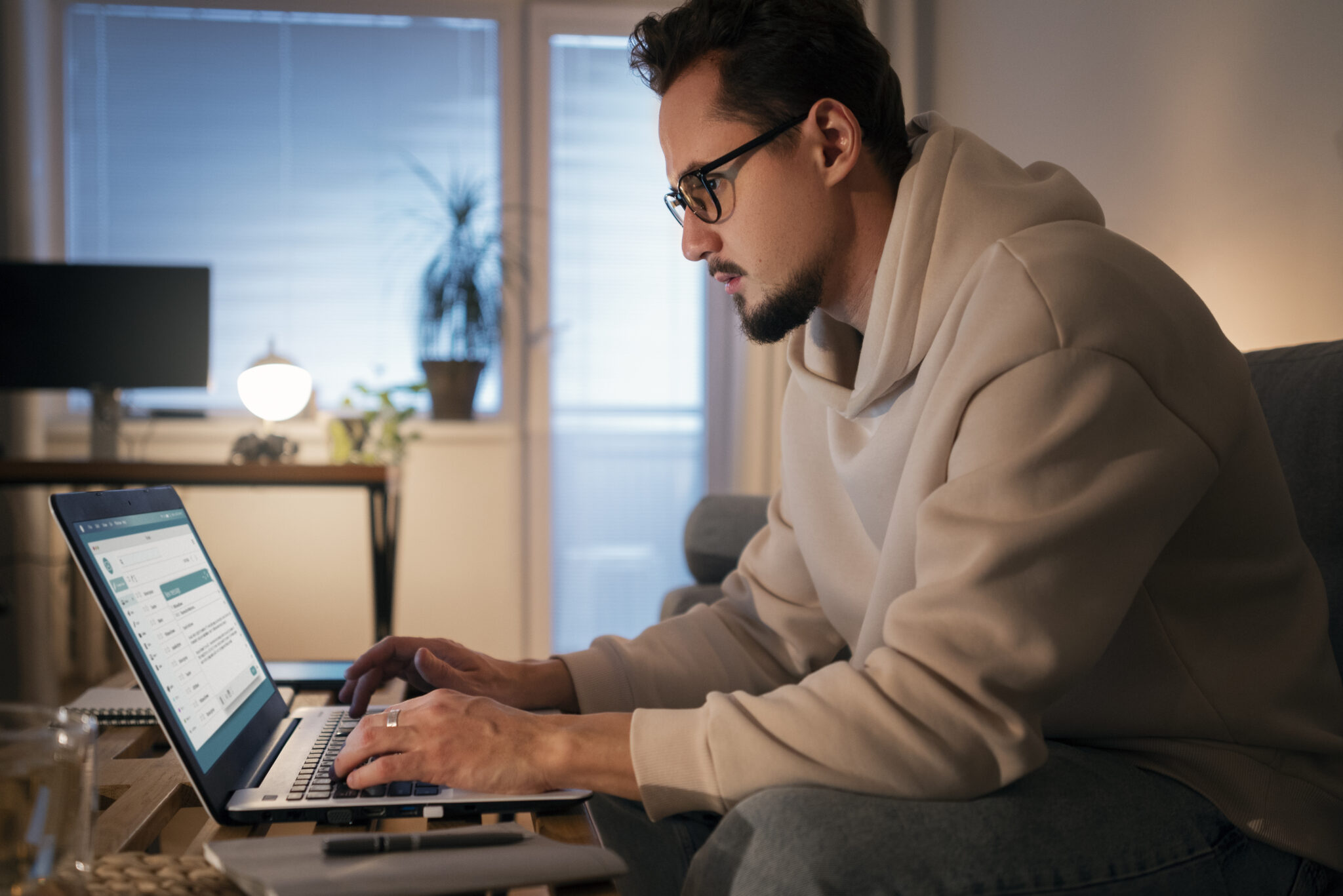 Side-view shot of a focused man with glasses and a beard wearing a light beige hoodie, sitting on a couch at home and typing on a laptop placed on a coffee table. Soft ambient lighting fills the room, showing a desk setup and window in the background.