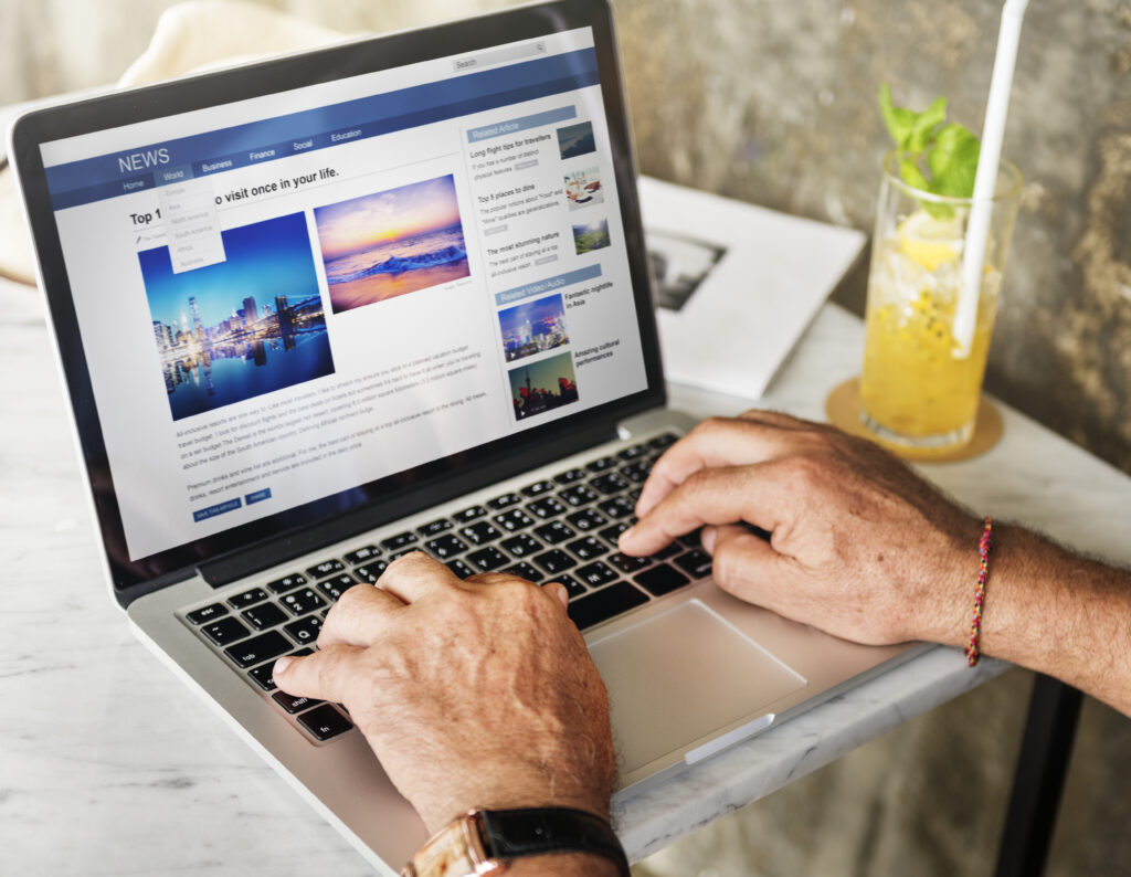Close-up of a person's hands typing on a silver laptop at a marble table next to a glass of iced beverage. The laptop screen displays an online news or travel blog layout featuring headlines, articles, and vibrant travel imagery.