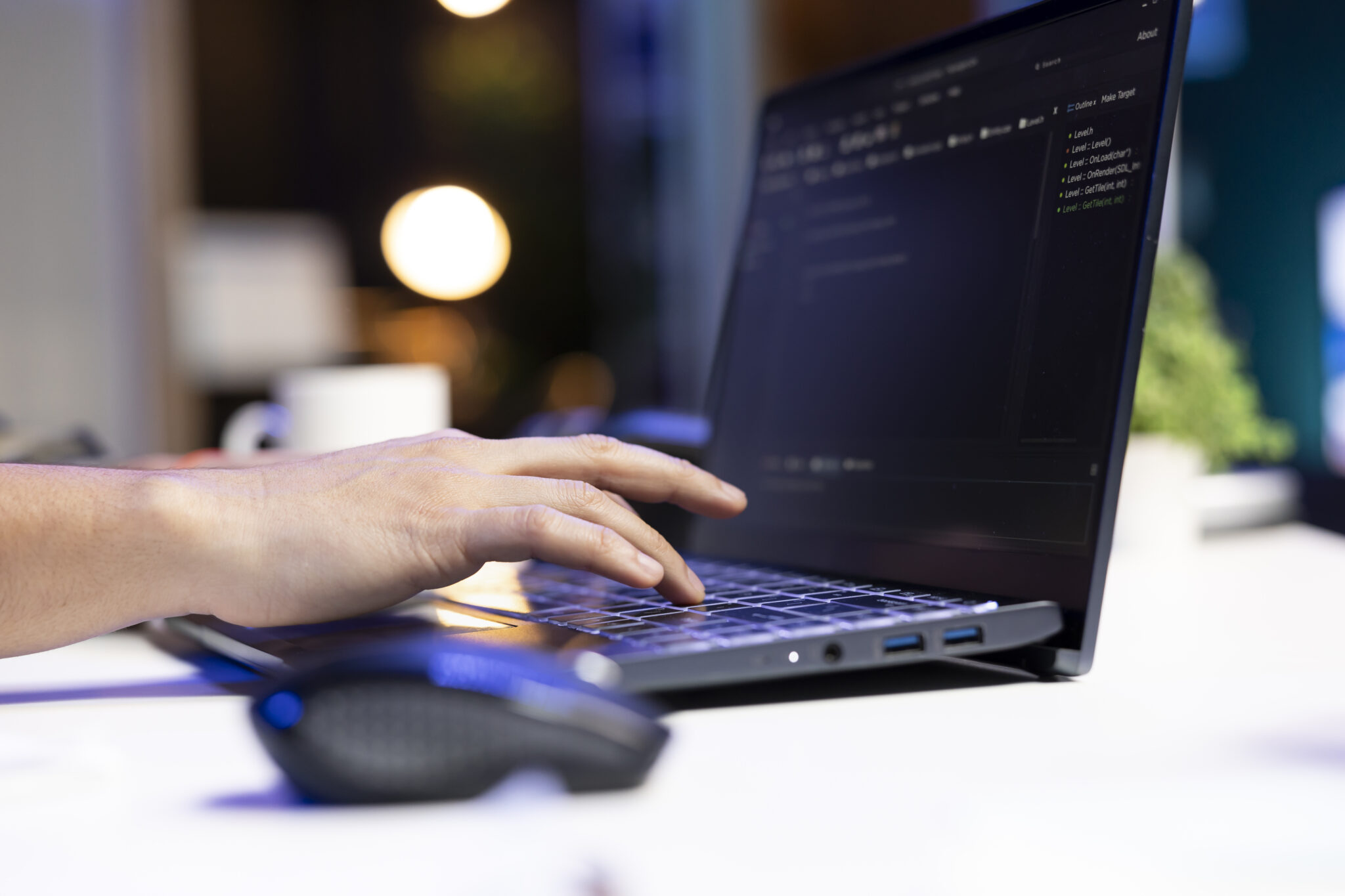 Side-angle view of a hand typing on a black laptop keyboard with illuminated keys on a desk at night. The computer monitor displays a dark-mode code editor or terminal interface, with a computer mouse blurred in the foreground and warm bokeh lighting in the background.